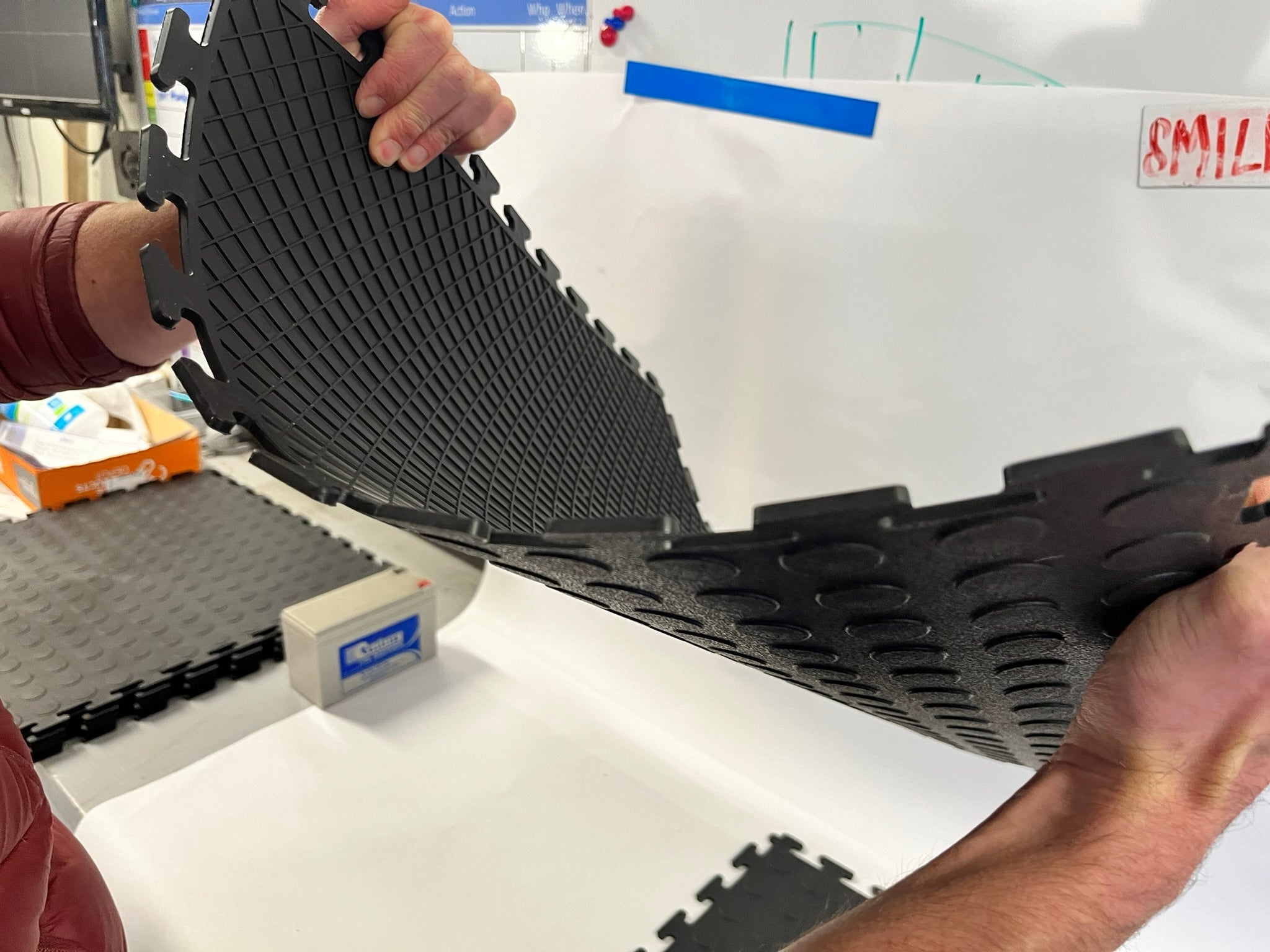 Person bending connected black garage floor tiles, showing their flexibility and grid-pattern underside.